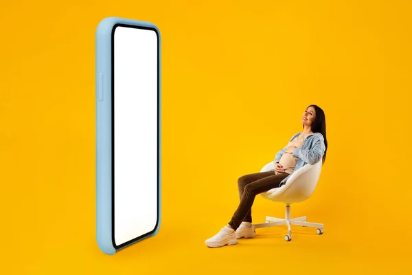 Happy pregnant woman sitting in chair near big blank smartphone with white screen, looking at copy space for online offer or mobile application design, mockup, full length