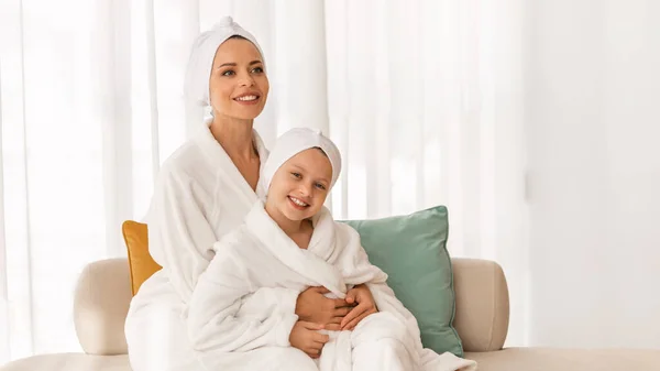 Portrait Of Beautiful Young Mom And Little Daughter Wearing Bathrobes And Towels On Head Relaxing On Couch At Spa, Happy Mother And Cute Female Child Having Beauty Day Together, Copy Space