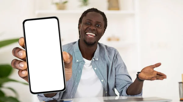 Cheerful Black Guy Sitting At Desk And Showing Big Blank Smartphone At Camera, Happy Young African American Man Holding Empty Cellphone, Recommending Mobile Application Or Website, Collage, Mockup