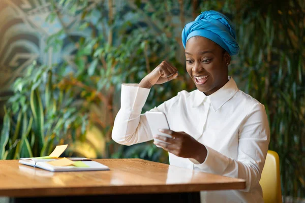 Happy excited young black businesswoman sitting at cafe, reading great news on cell phone, clenching fist and smiling, got email, positive answer from business partner, copy space