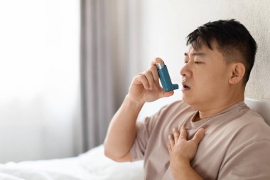 Sick unhealthy middle aged asian man in pajamas using inhaler and touching his chest, sitting in bed at bedroom alone, suffering from bronchial asthma attack, copy space