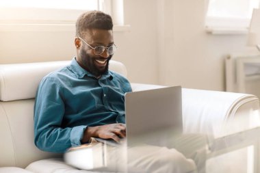 Excited black businessman sitting on sofa in office and using laptop, looking at display and taking part in video conference, free space. Freelancer working remotely