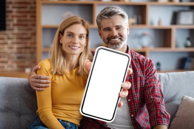 Happy Middle Aged Couple Sitting On Couch And Showing Blank Smartphone At Camera, Smiling Spouses Advertising New Mobile App Or Website, Recommending Online Offer, Creative Collage, Mockup