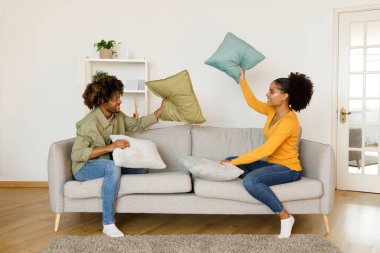 Joyful Black Couple Having Pillow Fight For Fun Sitting On Couch At Home. Young Spouses Throwing Pillows At Each Other Laughing And Flirting On Weekend. Happy Marriage And Relationship