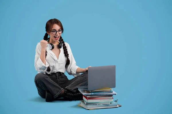 Satisfied teen european girl with pigtails in glasses with books and laptop rejoices to victory, make gesture of success and good scores, isolated on blue studio background. Study and education, ad