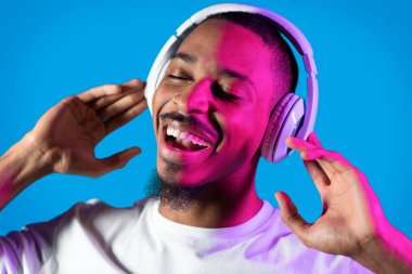 Joyful peaceful relaxed bearded young black guy in white listening to music with closed eyes and singing songs, using modern wireless headphones in neon light, blue background, closeup