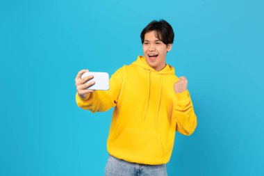 Joyful Asian Teenager Guy Holding Smartphone Gesturing Yes Winning Mobile Game Online Standing On Blue Background In Studio. Wow Application Advertisement Concept