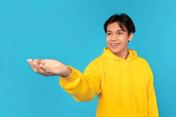 Look At This. Happy Chinese Teen Guy Stretching Hand To Camera Showing And Advertising Your Product Standing Over Blue Studio Background. Boy Holding Invisible Object.