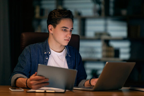Focused young handsome man in casual outfit employee sitting at workdesk, using laptop and digital pad, working at office in evening, copy space. Modern technologies in business
