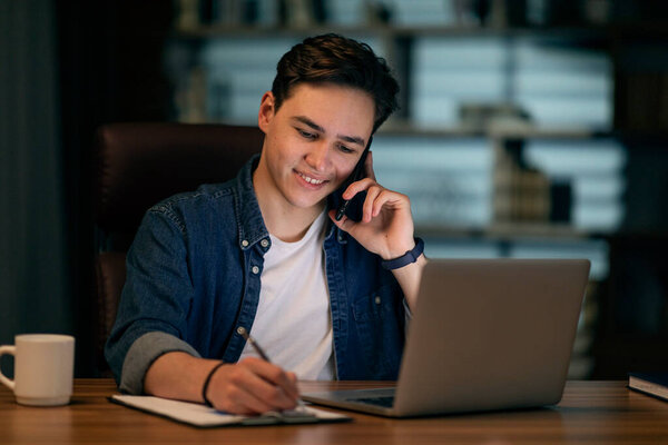 Happy young handsome guy in casual businessman working late at night, sitting at desk in front of computer, have phone call, taking notes, entrepreneur set business meeting, copy space