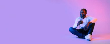 Cheerful handsome middle aged black man sitting on floor, using modern smartphone over neon studio background, gambling on Internet, chatting with ladies, looking at copy space, web-banner