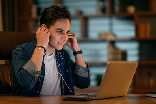 Young handsome cheerful man in casual outfit working for international company, guy employee sit at desk at dark office, using laptop, put earphones in, getting ready for online meeting, copy space