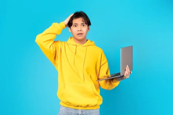 Oops, E-Learning Issue. Puzzled Japanese Teen Guy Holding Laptop And Touching Head Having Problem With PC Computer Standing On Blue Studio Background, Looking At Camera. Internet Connection Error