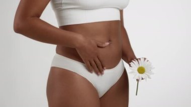 Female health concept. Close up shot of unrecognizable black woman in underwear caressing her bottom belly and holding camomile, white studio background, slow motion