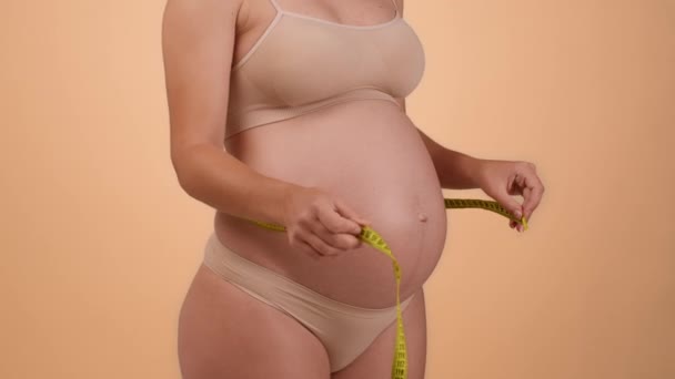 穿着内衣裤的健康孕妇的近照 用带子测量她的大肚子 检查她的生长 米色工作室背景 慢动作 — 图库视频影像