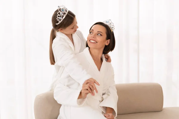 Cute Little Girl And Her Young Mom In Bathrobes And Toy Crowns Having Fun At Home, Portrait Of Happy Mother And Preteen Daughter Enjoying Beauty Spa Day Together, Looking At Each Other And Smiling