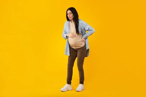 全长拍摄的怀孕年轻女子遭受情人背痛 孕妇触摸背部和腹部 站在黄色背景上 自由自在 — 图库照片