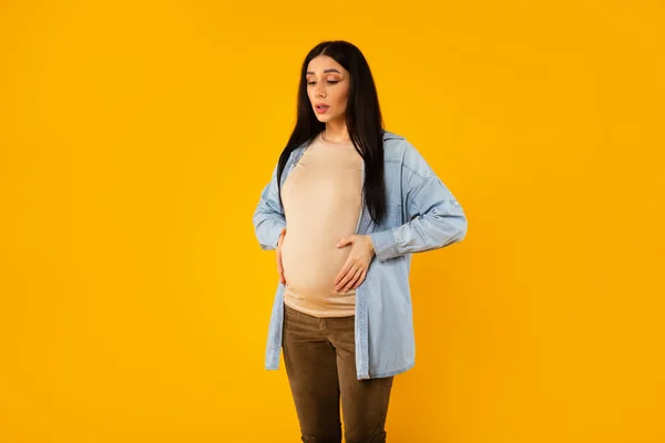 Pregnant lady suffering from pain, having painful spasm and touching belly, doing breathing exercises over yellow background. Pregnancy and healthcare problems