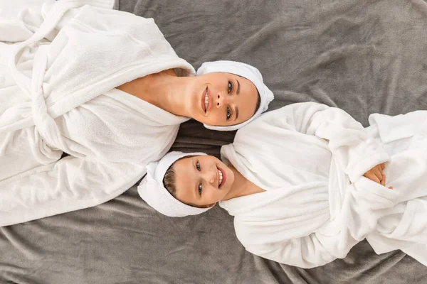 Spa And Beauty Concept. Mom And Little Daughter In Bathrobes And Towels On Head Relaxing On Bed Together, Happy Young Mother And Preteen Female Child Resting After Bath, Enjoying Skincare Treatments