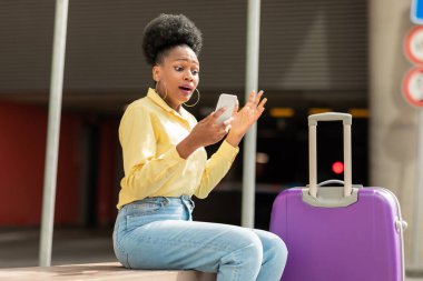 Shocked Black Female Tourist Using Cellphone Having Problem Travel Application Sitting With Suitcase Outdoors. Lady Having Issues With Mobile Connection Traveling Abroad. Bad Roaming Service