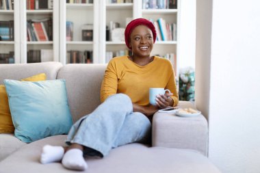 Cheerful happy attractive millennial black woman in homewear and african turban chilling alone on couch at home, drinking coffee, looking at copy space and smiling. Lifestyle, relaxation, weekend