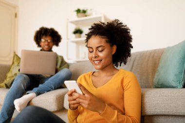 Happy African American Woman Using Cellphone Texting While Her Husband Browsing Internet On Laptop Computer Sitting In Living Room At Home. Spouses Using Gadgets On Weekend. Selective Focus