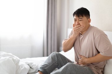 Sick middle eastern korean man in pajamas sitting in bed at home, covering mouth, touching belly, asian male suffering from nausea in the morning, copy space. Food poisoning, vomiting, indigestion