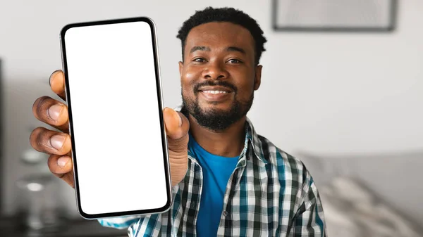 Mobile Ad. Happy Black Man Demonstrating Big Blank Smartphone With White Screen At Camera While Sitting On Couch At Home, Young African American Male Advertising New Mobile App, Collage, Mockup