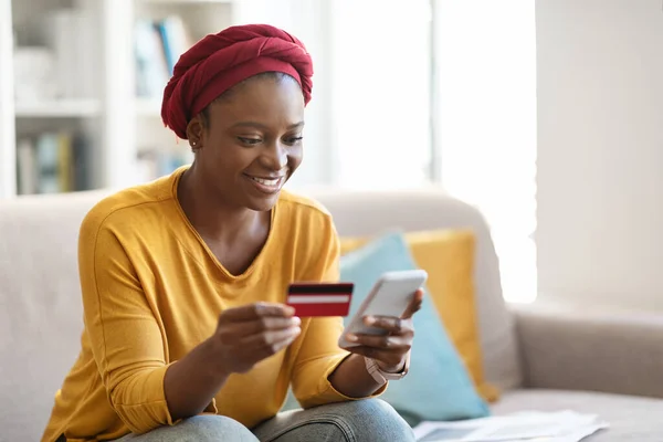 Online shopping, e-commerce, retail concept Cheerful pretty young black lady wearing african turban and casual outfit sitting on couch at home, holding credit card and smartphone, copy space