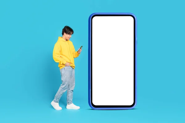 Chinese Teenager Guy Using Smartphone Walking Near Large Cellphone With Empty Screen On Blue Background In Studio. Boy Texting And Using Social Media App Near Big Gadget. Mockup, Full Length