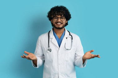 Cheerful handsome young indian man in workwear and eyeglasses doctor with stethoscope smiling and gesturing welcome over blue studio background, copy space. Healthcare, medical staff concept