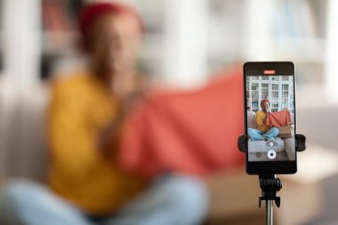 Selective focus on cell phone set on tripod recording pretty young african american woman fashion blogger influencer showing brand new cloth, unpacking delivery, home interior, blurred background