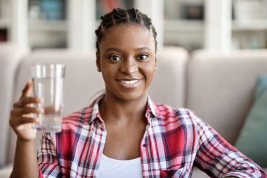 Pretty cheerful young african american woman in casual holding glass of fresh mineral water and smiling at camera, home interior, copy space. Hydration, healthy lifestyle concept