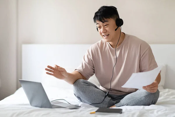 Cheerful friendly middle eastern Asian man working from home, sitting on bed wearing pajamas, using pc laptop and headset, holding papers, have online meeting with colleagues, copy space