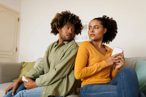 Jealousy In Relationship. Displeased Black Husband Peeking At Wifes Phone While She Texting Suspecting Affair Sitting On Sofa At Home. Infidelity And Marriage Problems Concept