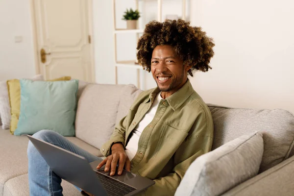 Happy Black Freelancer Man Using Laptop Computer Smiling Looking At Camera Sitting On Sofa, Working Remotely Online At Home. Successful Freelance And Distance Job Concept