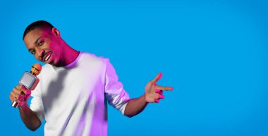Cheerful handsome charismatic young black guy in white entertainer or singer posing with microphone in his hand over blue studio background in neon light, panorama with copy space