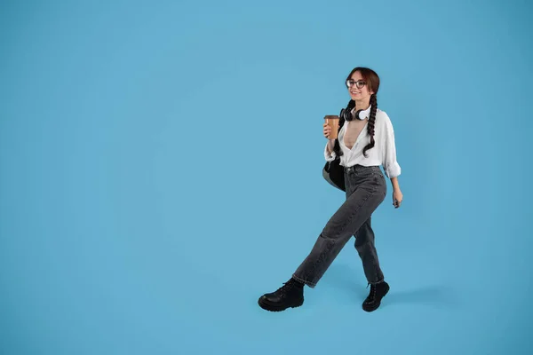 Smiling teenage european girl with pigtails in glasses, headphones with backpack drink cup of coffee takeaway and walk, isolated on blue studio background. Ad and offer, break at study, education