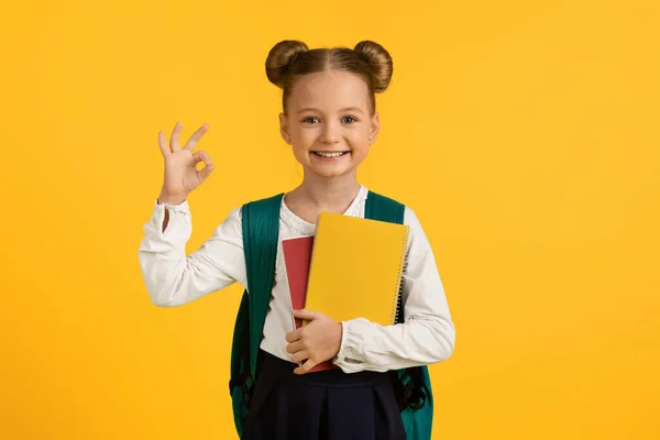 Cute Little Schoolgirl With Workbook In Hands Showing Ok Gesture, Happy Female Pupil With Backpack Gesturing Okay, Recommending Education Program While Standing Isolated Over Yellow Background