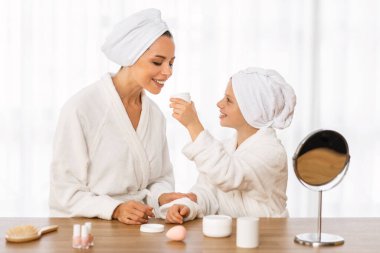 Anne ve küçük kızı bornozlarını denerken evde güzellik rutini yapıyorlar, mutlu genç anne ve kadın çocuk kavanozu nemlendirici kremle kontrol ediyorlar, cilt ürünlerinin tadını çıkarıyorlar.