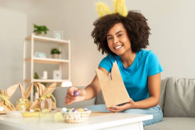 Paskalyayı evde kutlayan mutlu siyahi kadın portresi, tatil hediyeleri hazırlayan, tavşan kulaklı neşeli Afro-Amerikan kadın, el yapımı kağıt torbalara çikolatalı yumurta koyuyor.