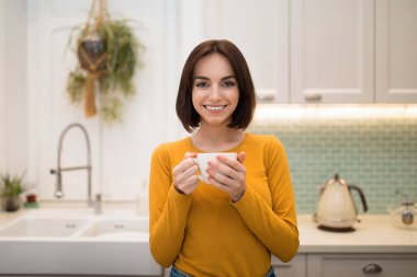 Güzel esmer, neşeli genç bayan evde bitki çayı içiyor, modern, rahat mutfakta duruyor, kahve fincanı tutuyor ve kameraya gülümsüyor, kadın işten sonra dinleniyor.