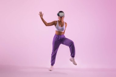 Fit Afro-Amerikan bayan dans ediyor ve sanal gerçeklik gözlüğü takarken metaevrende eğleniyor, pembe stüdyo ışıklarında 3 boyutlu teknolojiyi keşfediyor.