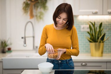Rahat kıyafetler içinde, evde sağlıklı kahvaltı hazırlayan, ev yapımı tahıl gevreğiyle ev yapımı kahvaltı yapan, rahat görünümlü genç bir kadın. Diyet, beslenme, detoks, sağlıklı yaşam tarzı konsepti.