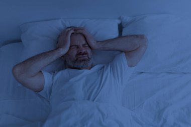 Mutsuz, gri sakallı, pijama giyen, kafasına dokunan, geceleri baş ağrısı çeken ve migren ağrıları çeken olgun bir adamın görüntüsü.