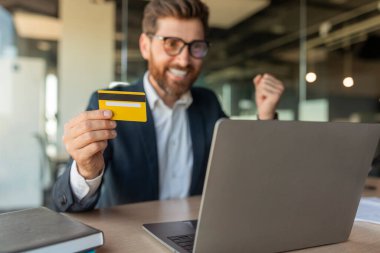 Çok mutlu bir iş adamı, ofisin içindeki masada oturan dizüstü bilgisayarın önünde kredi kartı tutarak başarı jesti yapıyor. Finansal anlaşma, iş ve iş, online alışveriş