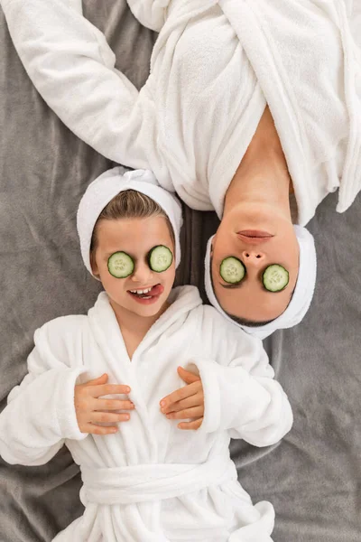 Mother and daughter doing beauty treatment together at home, applying cucumber slices on eyes, top view shot of happy young mom and female child in white bathrobes relaxing on bed after spa day