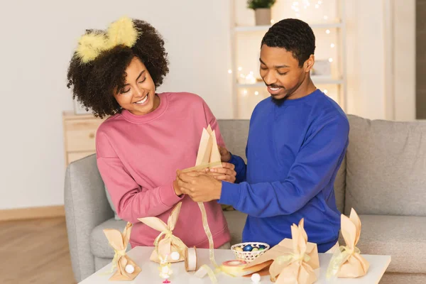 黒若いカップル作るイースター装飾で自宅一緒に 幸せなアフリカ系アメリカ人配偶者準備祭ギフト包装 包装クラフトバッグでリボンと笑顔 女性を身に着けているバニーの耳 — ストック写真