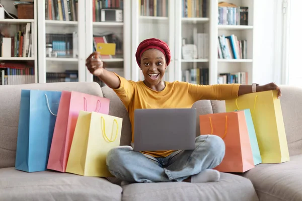幸せな赤いターバンのお店ではかなり若い黒人女性の興奮カラフルなショッピングバッグの多くは 自宅でラップトップを使用して クレジットカードを示すソファに座っている 電子商取引 オンラインショッピング — ストック写真