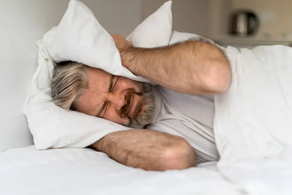 I Cant Sleep. Portrait of irritated middle aged man lying in bed and covering ears with pillows, hearing and suffering from too loud sound or snoring, tired of noisy neighbors. copy space